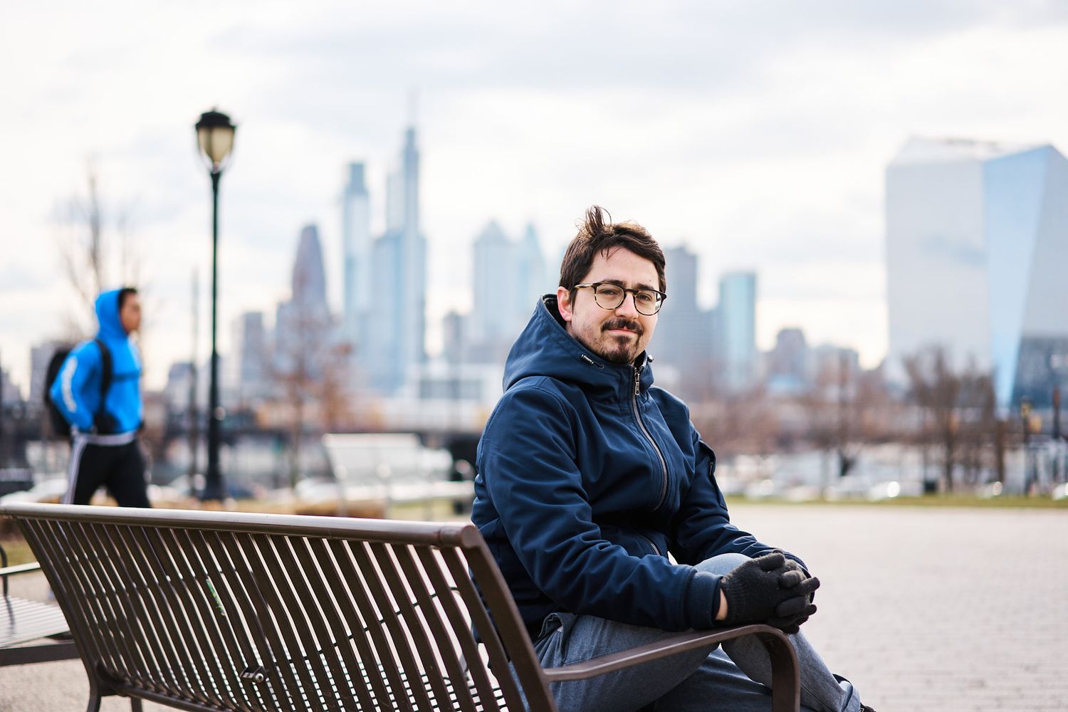Man in blue jacket sitting on park bench with Philadelphia skyline in background on cloudy day.