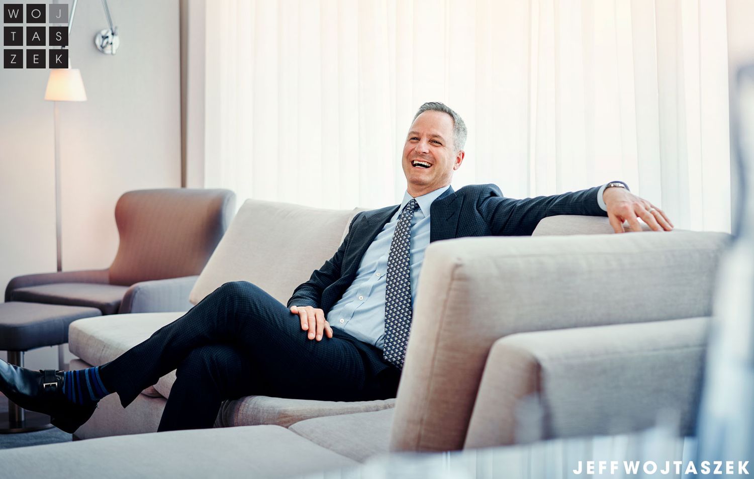 Smiling businessman in suit relaxing on modern sofa, laughing in stylish lounge setting.