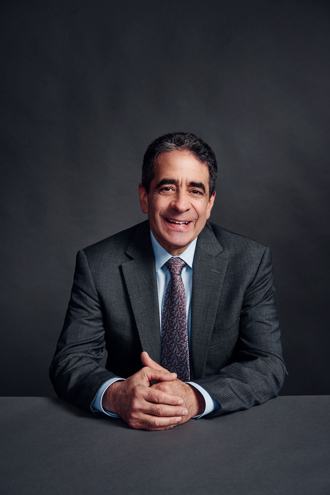 Smiling businessman in gray suit and tie sitting at table with hands clasped against dark background.