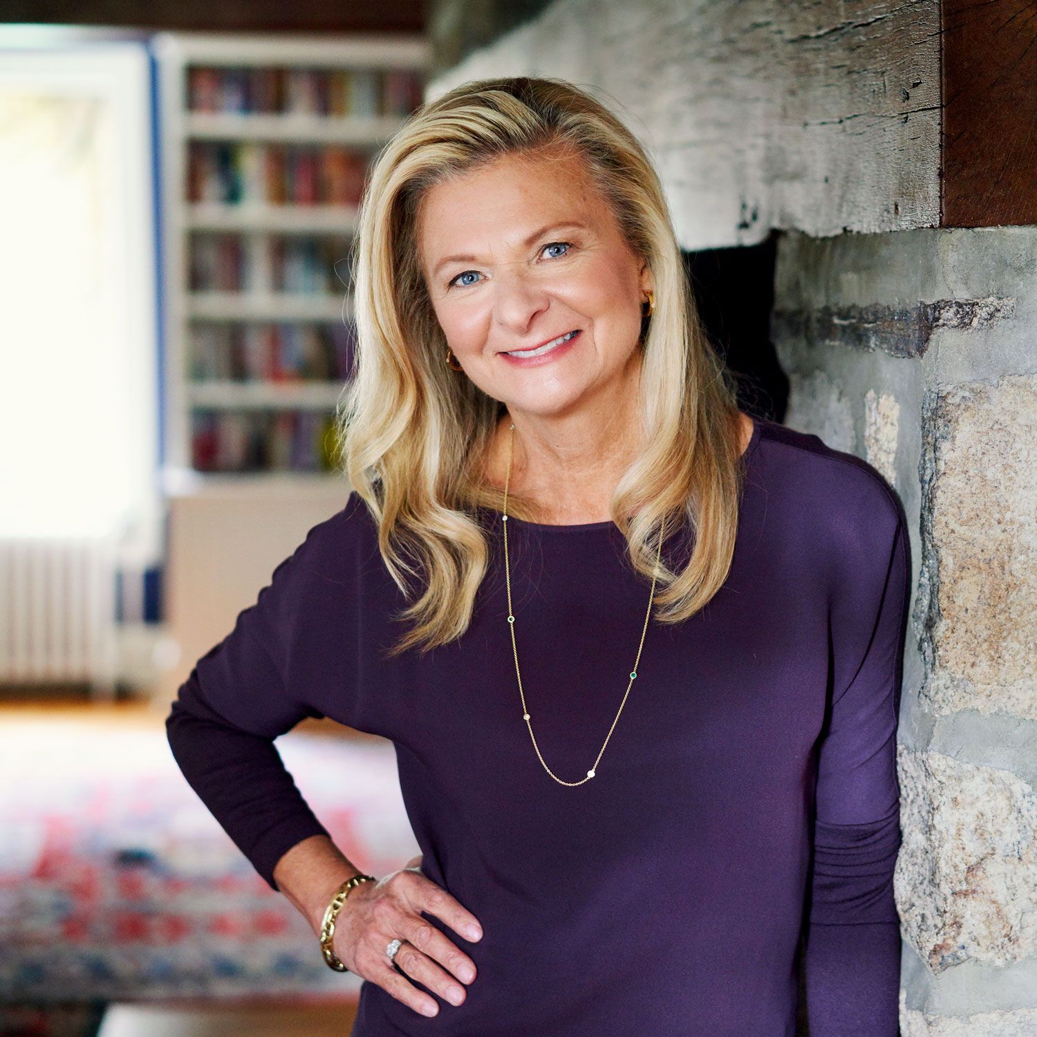 Smiling blonde woman in purple top posing beside a stone fireplace indoors.