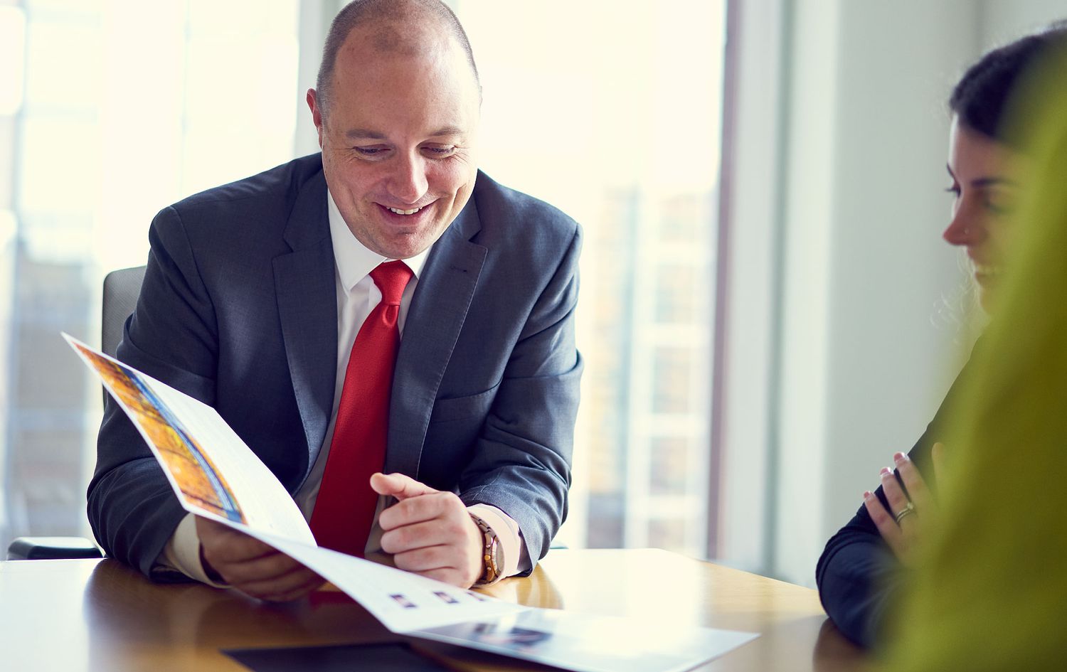 Smiling businessman in suit and red tie reviewing colorful reports with a colleague in bright office.