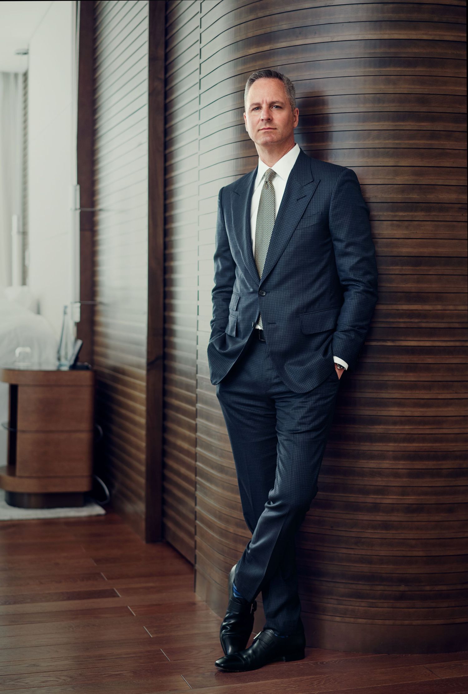 Professional man in dark suit and tie leaning against wooden wall in modern interior setting.