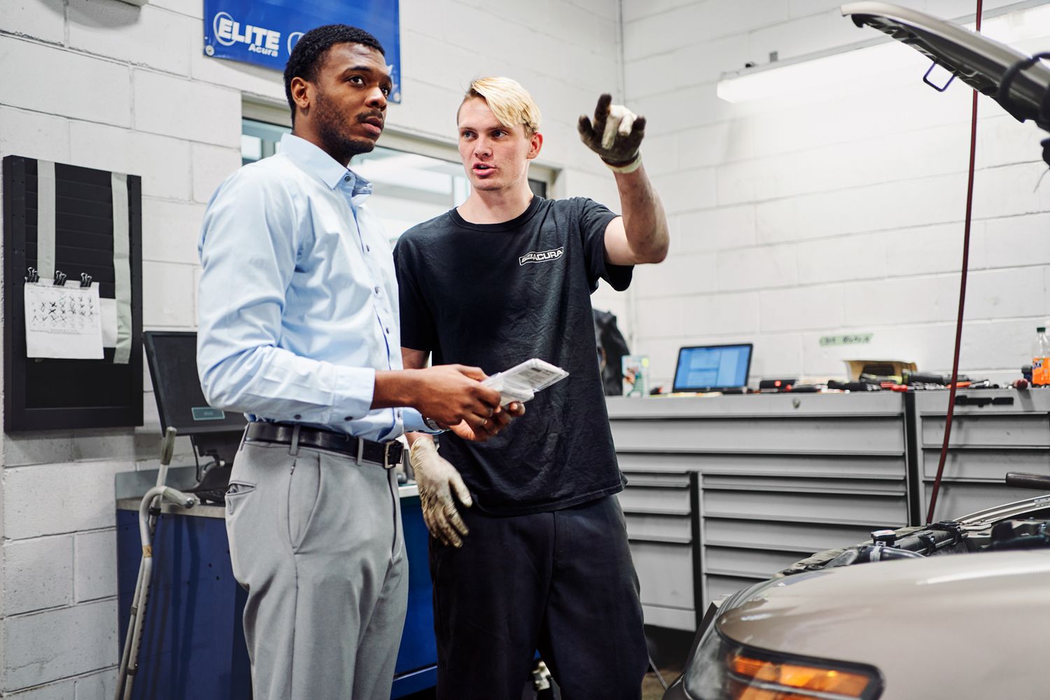 Two auto mechanics discussing vehicle repair in a professional garage, one holding a tablet and the other wearing work gloves.