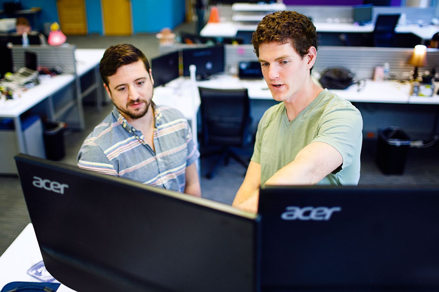 Two male colleagues collaborating at dual Acer monitors in a modern open-plan office workspace.