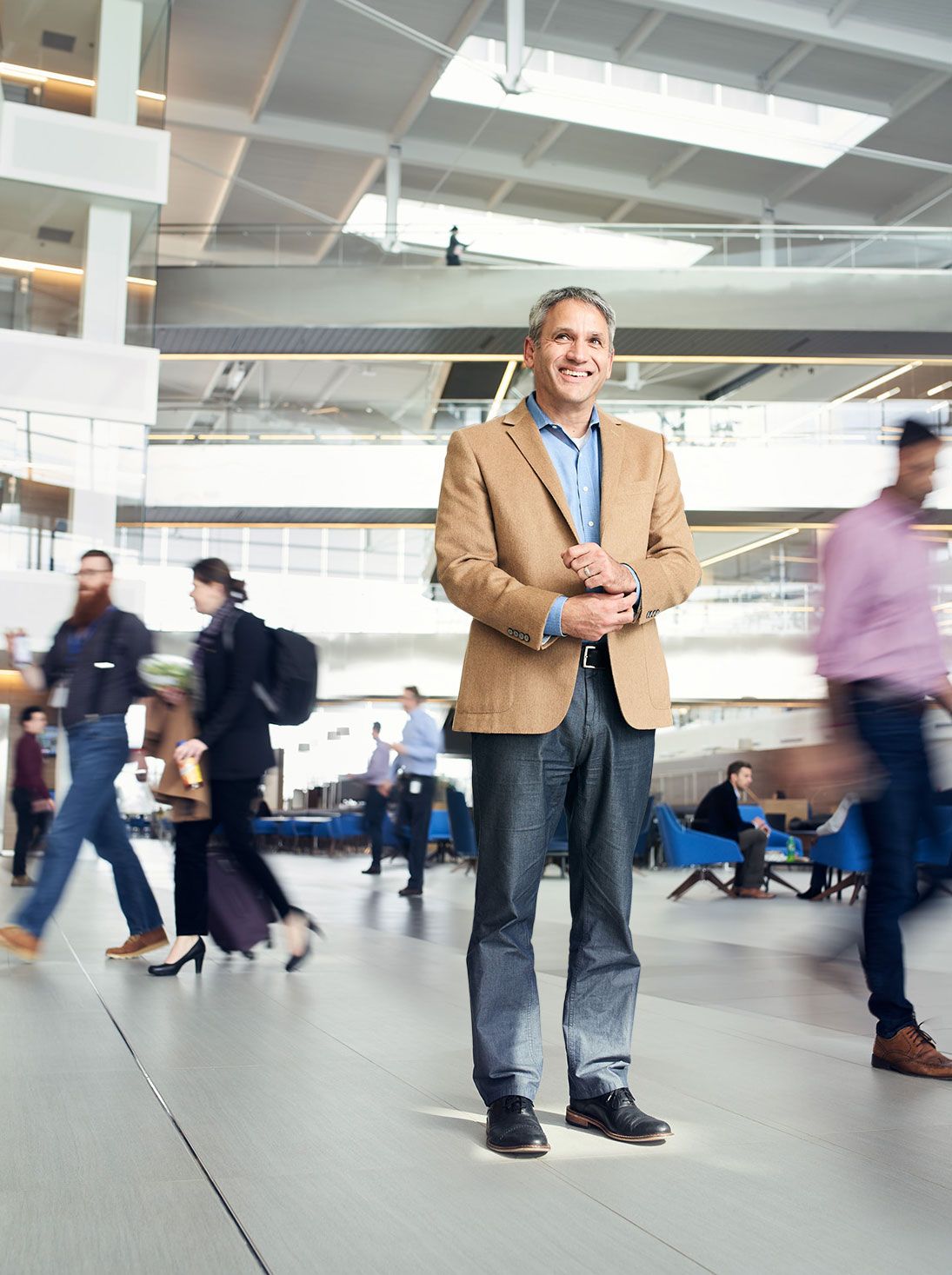 Confident businessman in tan blazer standing in busy modern office atrium with blurred crowd.
