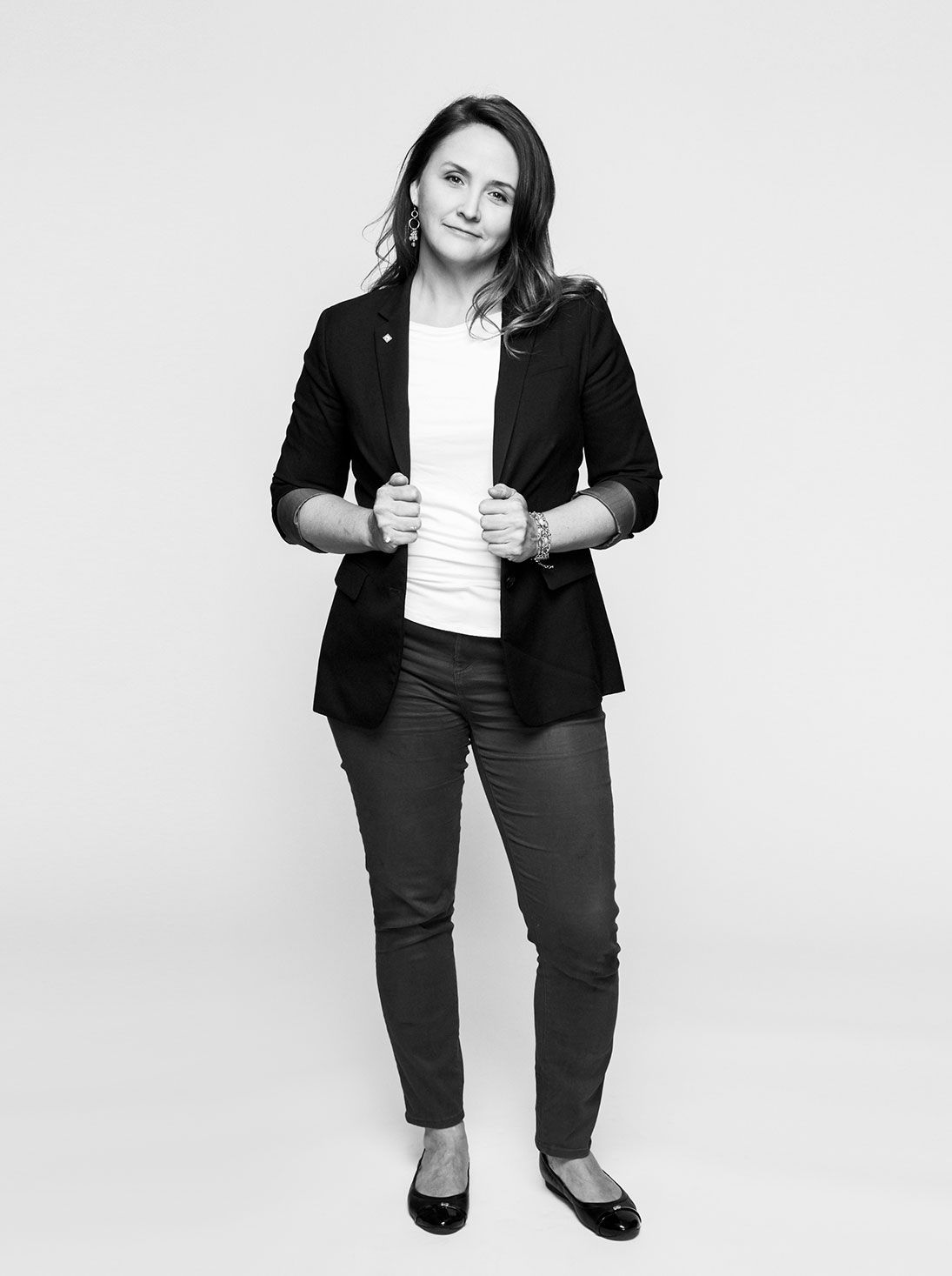 Confident woman in black blazer and grey pants poses in a black and white full-length portrait.