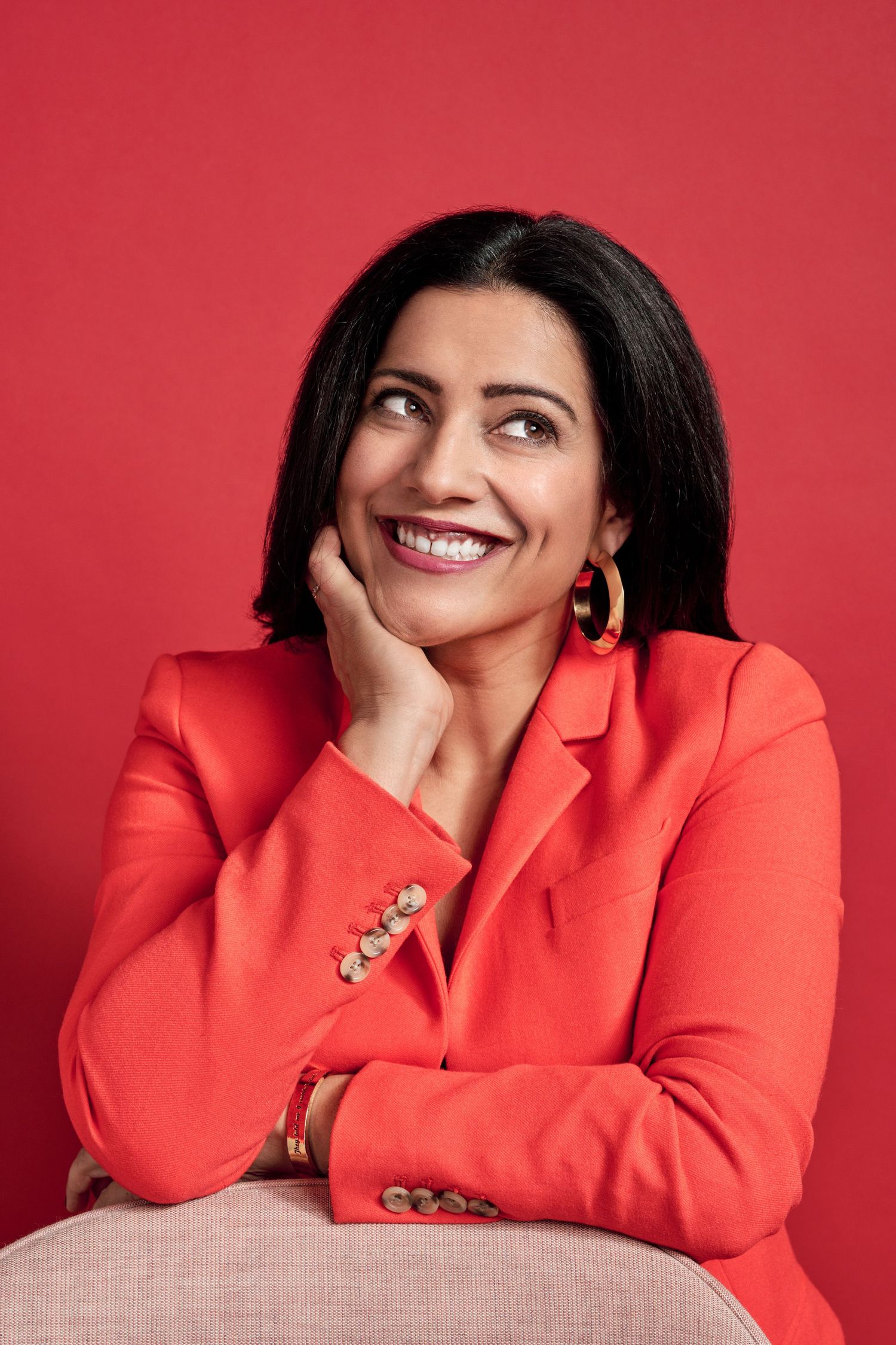 Smiling woman in red blazer and hoop earrings poses against red background with hand on chin.