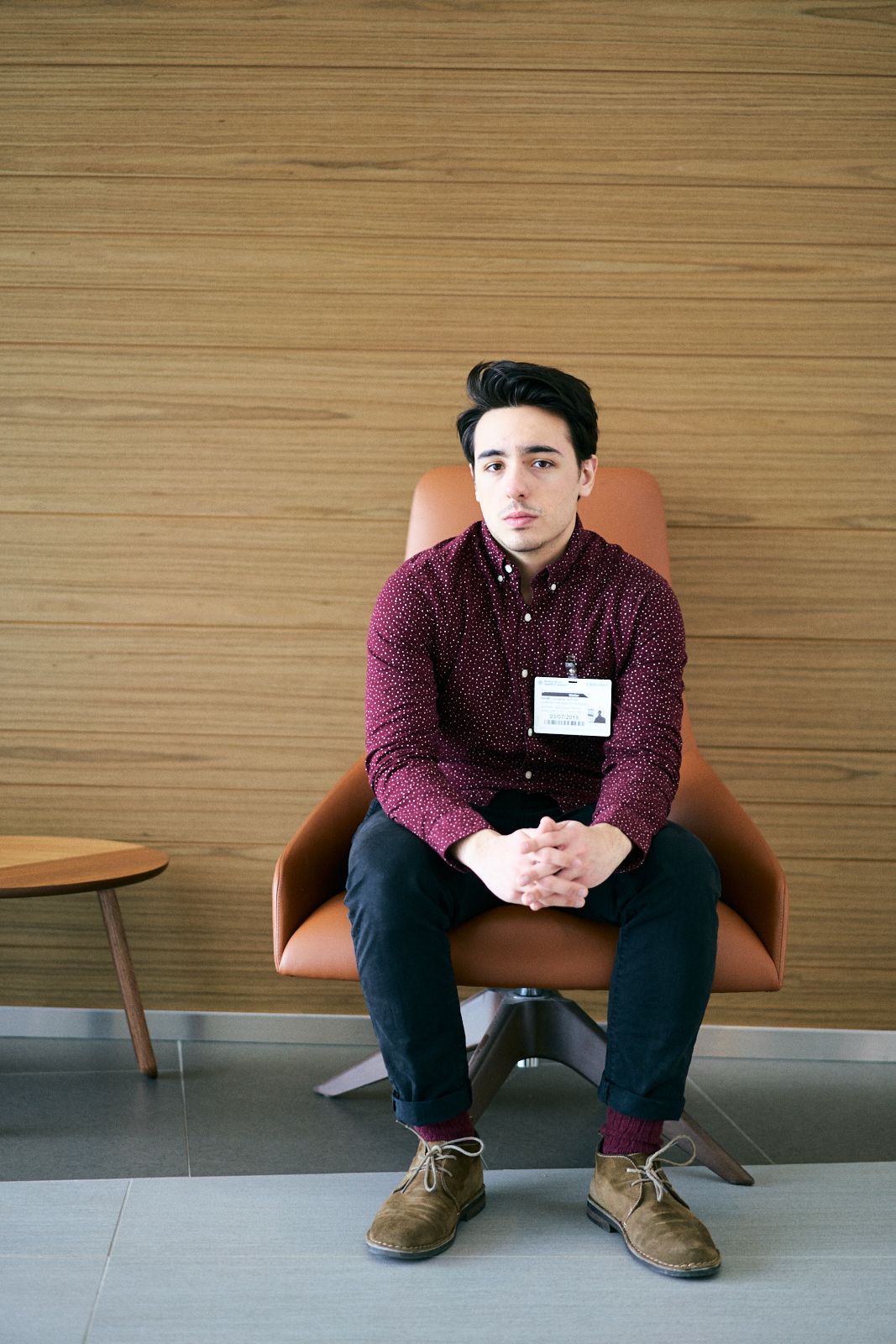 Young man in burgundy shirt sitting in orange leather chair against wood panel wall wearing a name badge.