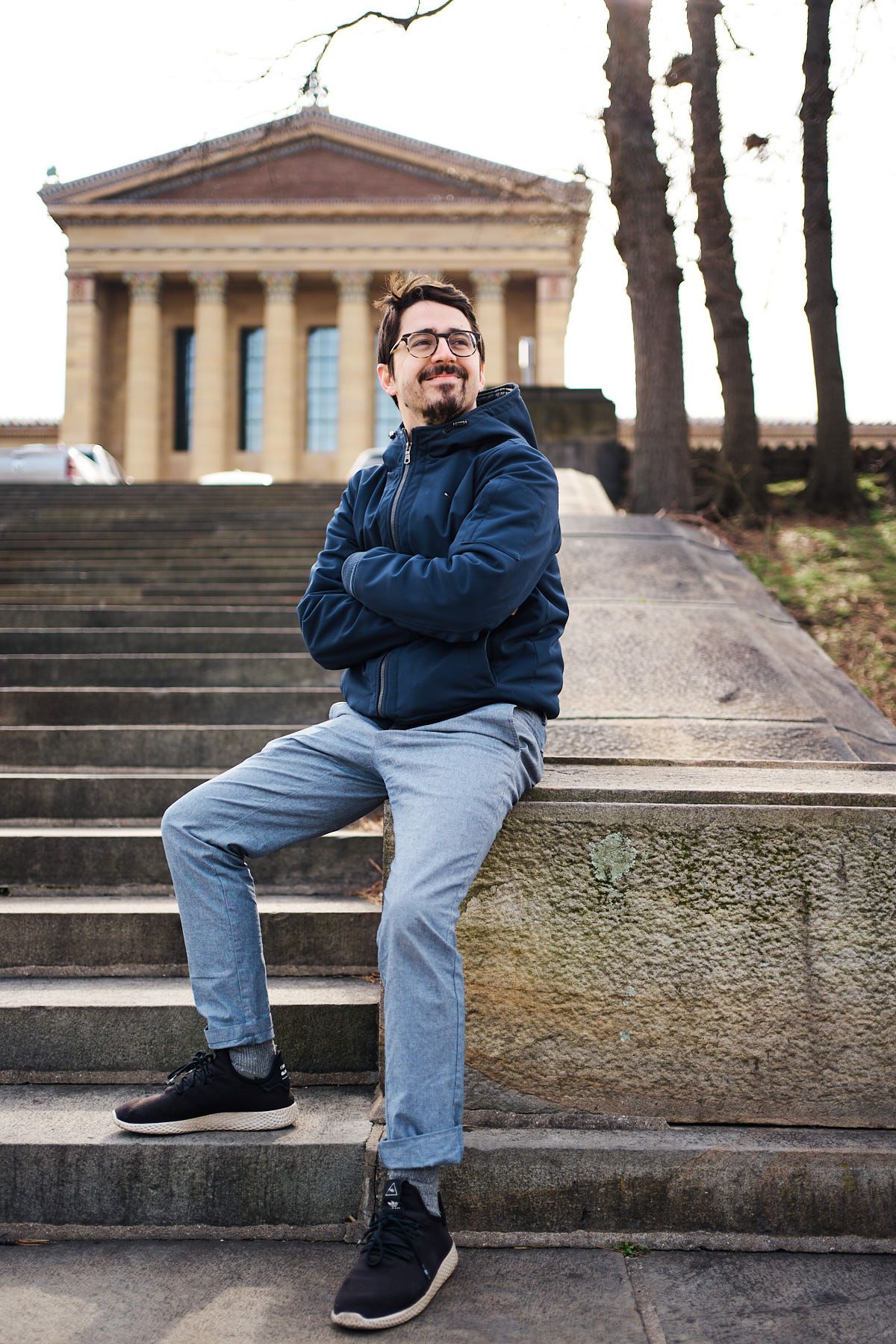 Man in blue jacket sitting on steps outside a classical building with columns, smiling with arms crossed.