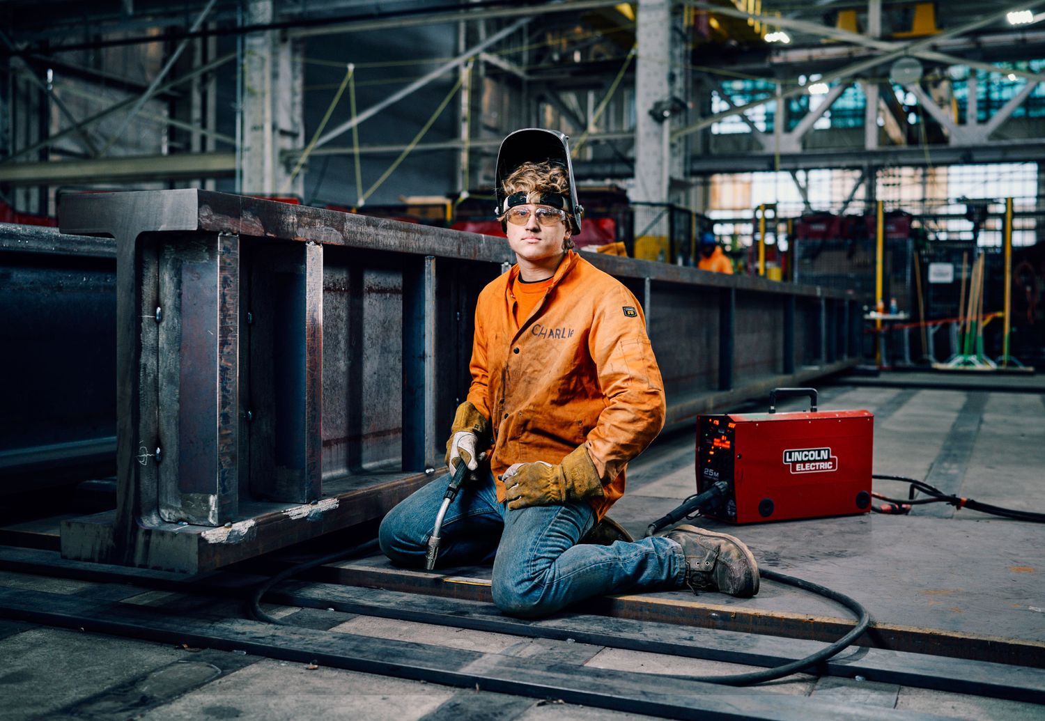 Welder in orange jacket and safety gear kneeling beside steel beams in an industrial warehouse.