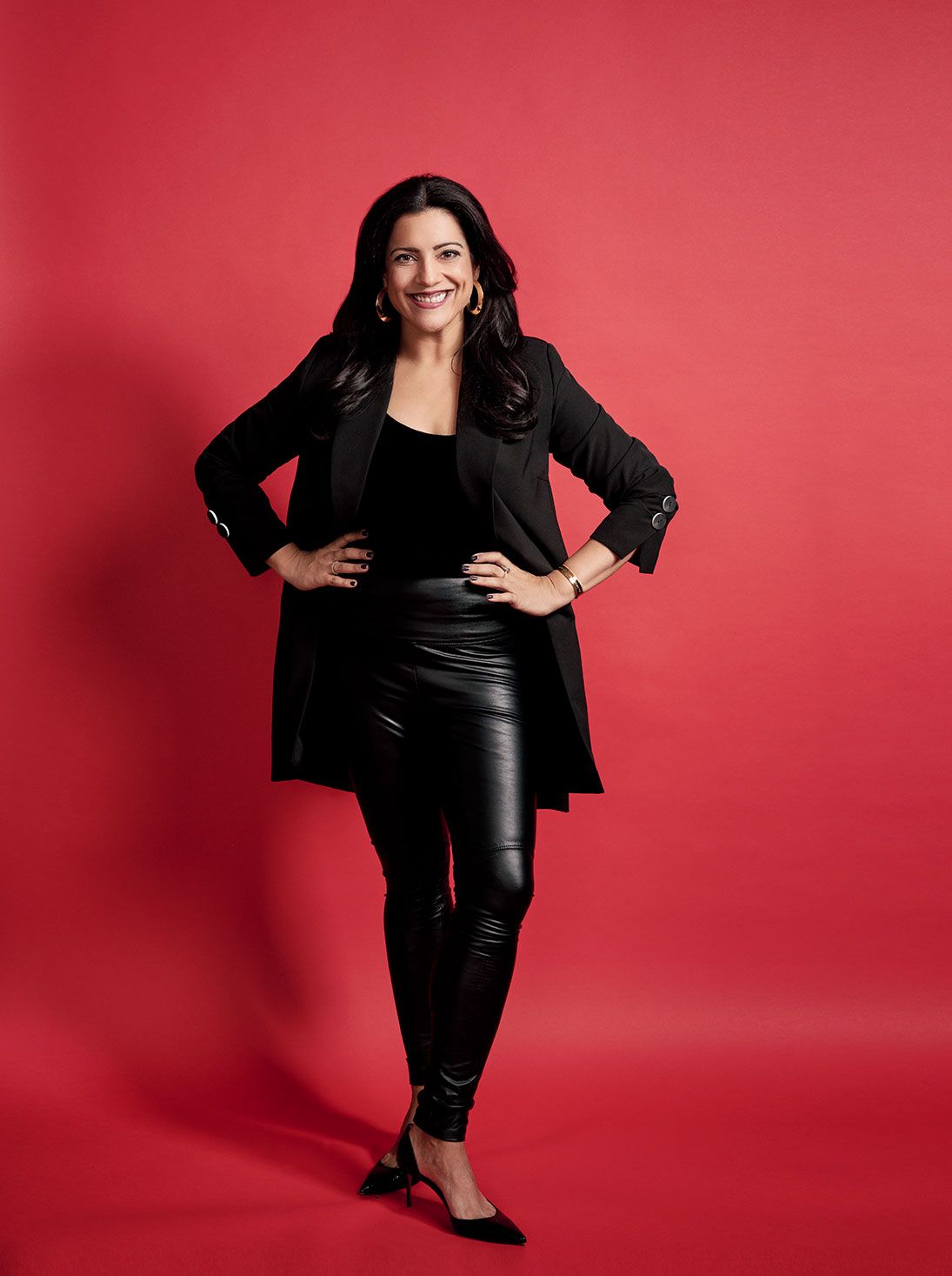 Confident woman in black blazer and leather pants posing against vibrant red background.