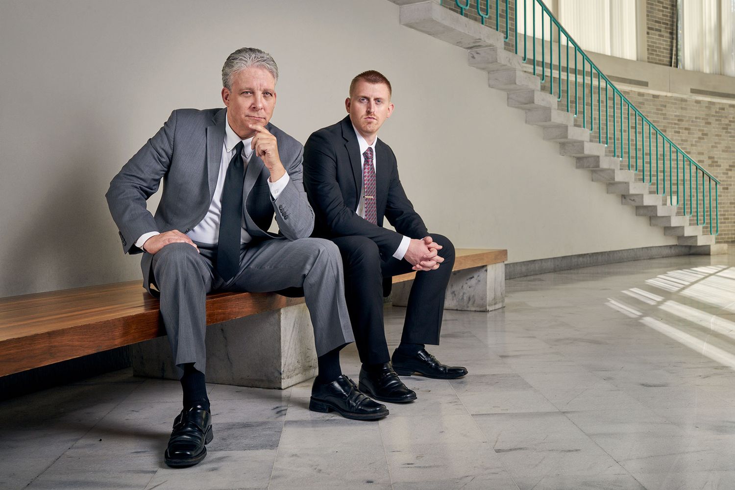 Two businessmen in suits sitting on bench in modern marble building interior.