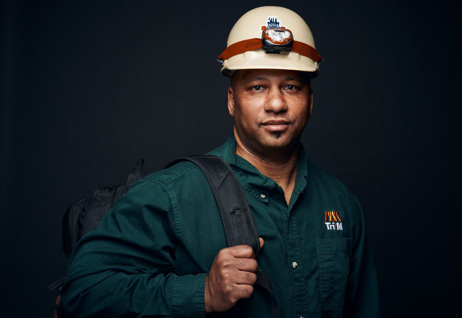Professional worker in hard hat and headlamp with backpack poses confidently against dark background.