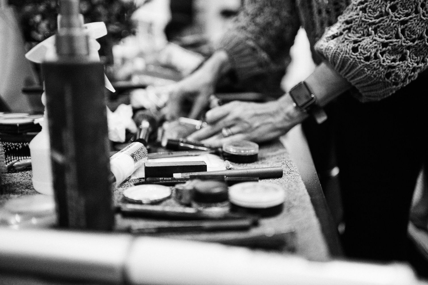 Black and white photo of hands working at a makeup artist station with brushes and cosmetics on a table.