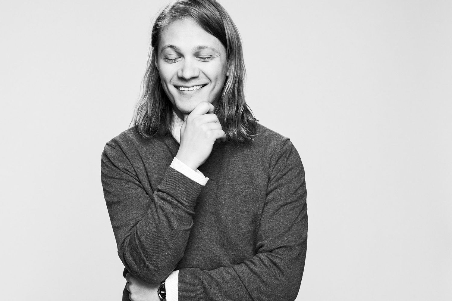 Black and white portrait of a young man with long hair smiling thoughtfully with hand on chin.