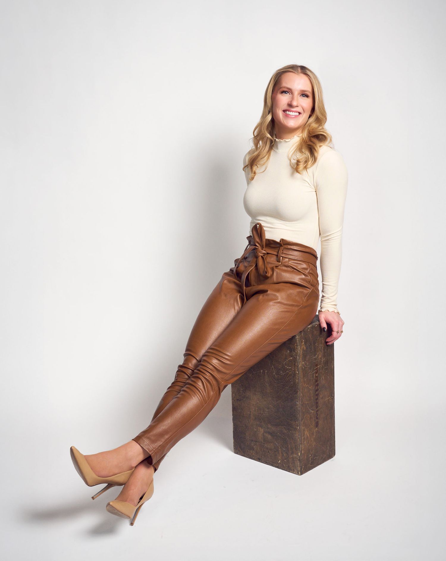 Smiling blonde woman posing in brown leather pants and cream turtleneck sweater seated on wooden block.