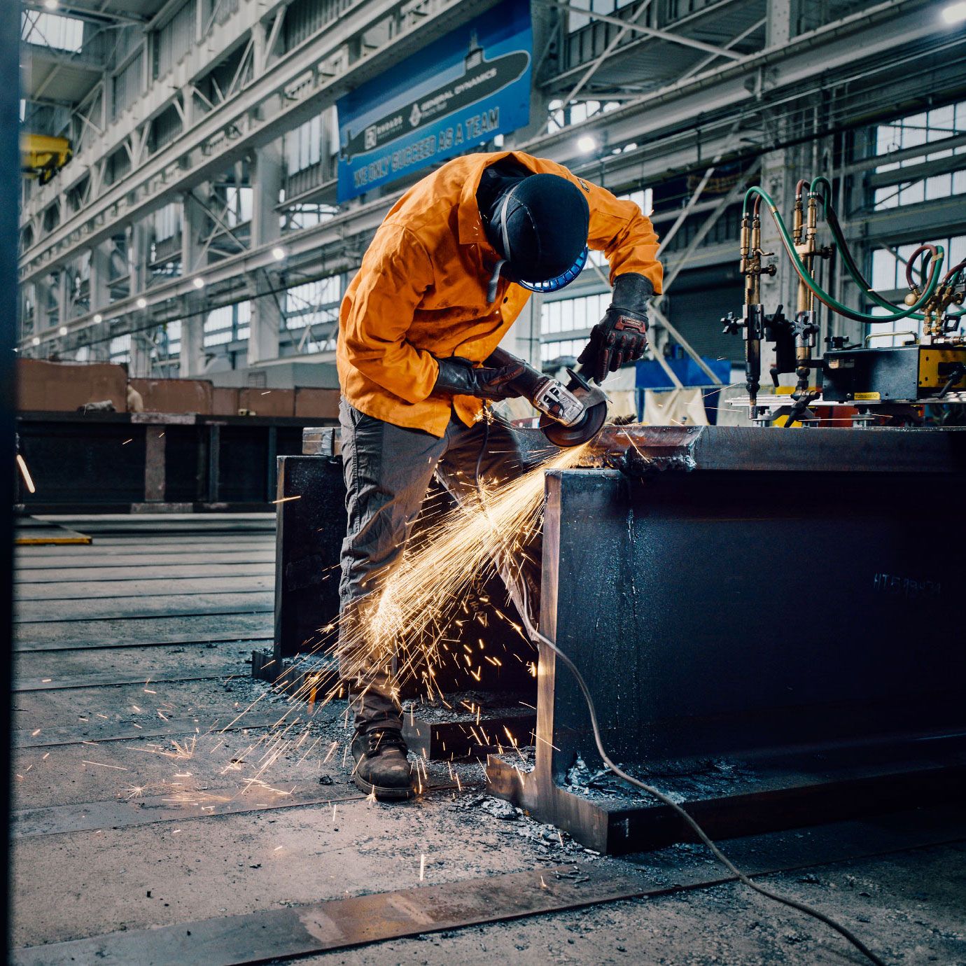 Worker in orange jacket grinding metal with angle grinder, sparks flying in industrial factory setting.