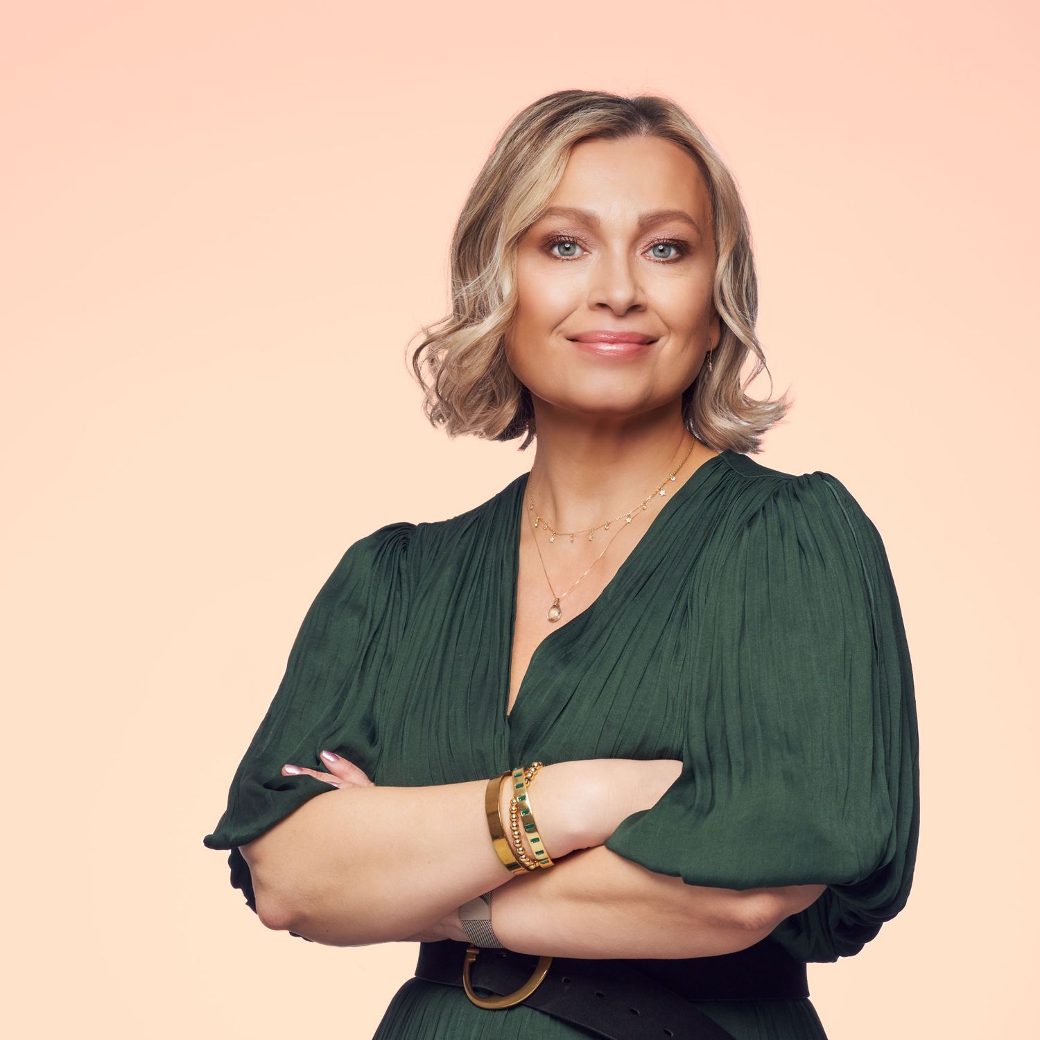 Confident blonde woman in green blouse with arms crossed against peach background.