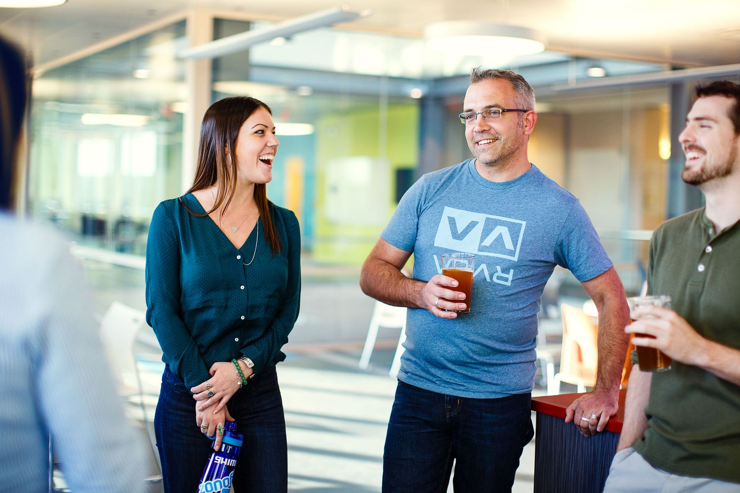 Three colleagues laugh and chat while holding drinks in a bright modern office during a casual work social event.
