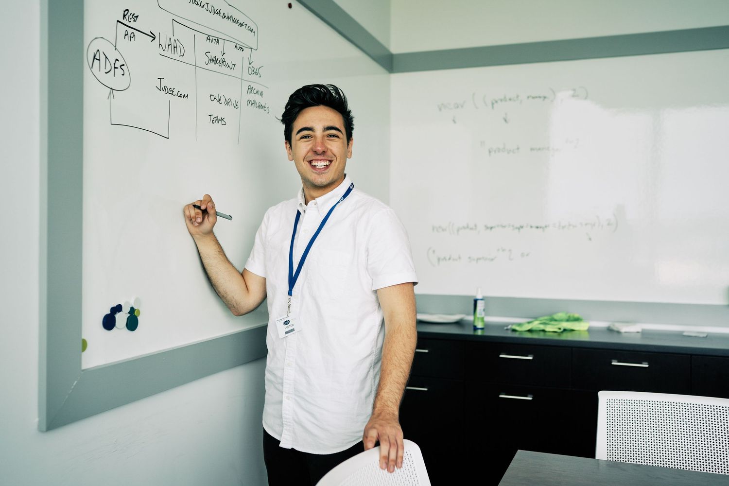 Smiling man in white shirt with lanyard stands at whiteboard in modern office, holding marker and paper.