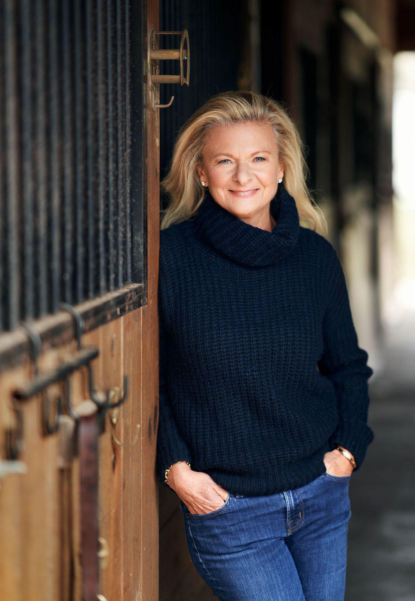 Smiling blonde woman in navy turtleneck sweater leans against a wooden stable door.