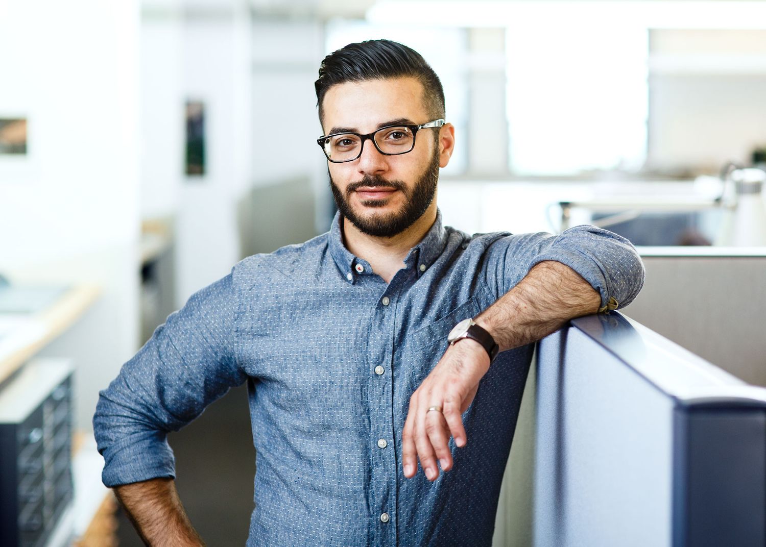 Professional man with glasses and beard leaning on office cubicle in modern workplace.