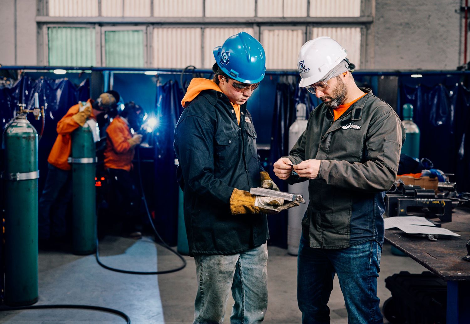 Two workers in hard hats reviewing documents near welding equipment in industrial workshop.