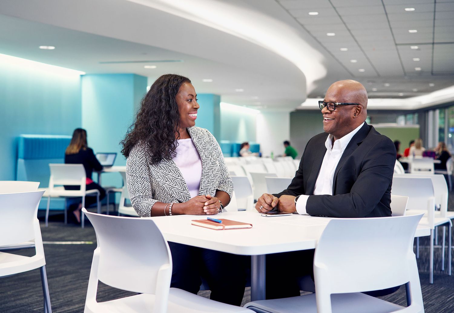 Two business professionals share a friendly conversation at a white table in a modern office cafe.