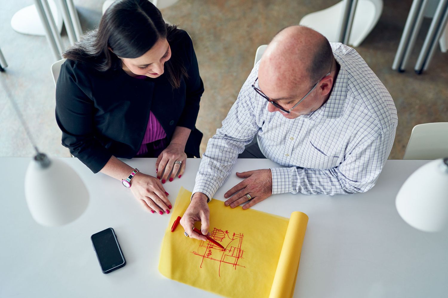 Overhead view of two colleagues reviewing architectural blueprints on yellow paper at a white office table.