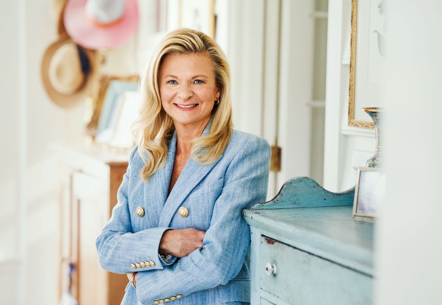 Smiling blonde woman in blue blazer leaning against a dresser indoors.