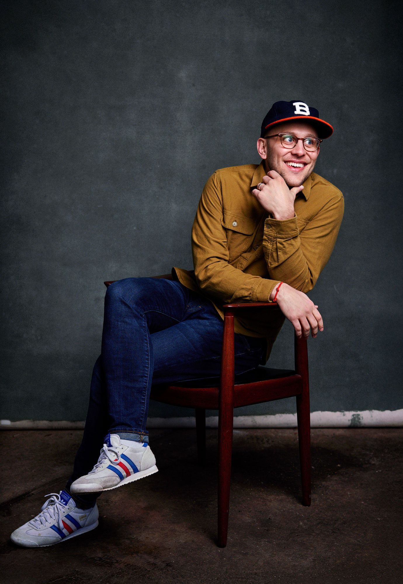 Young man smiling, seated on wooden chair, wearing brown shirt, blue jeans, sneakers, and a baseball cap with glasses.