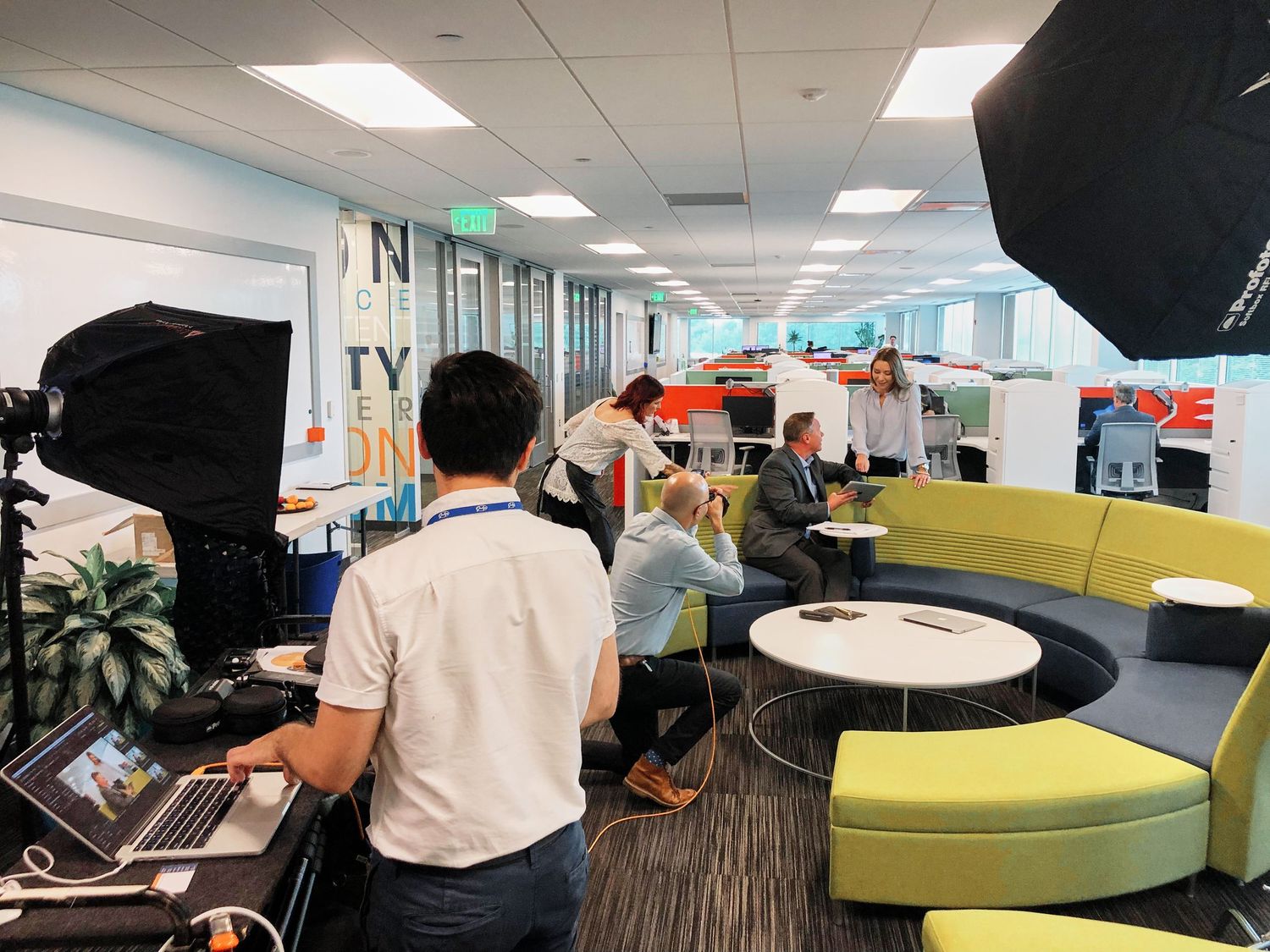 A professional video production crew films an interview in a modern open-plan office with colorful furniture.