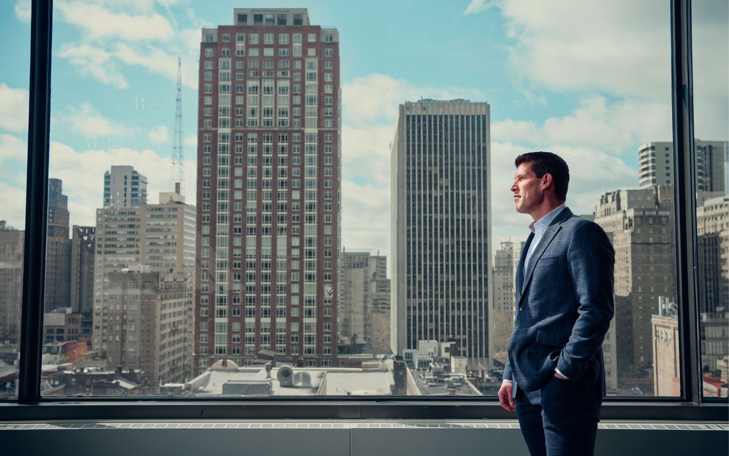 Young businessman in blue suit gazing at city skyline through large office window.