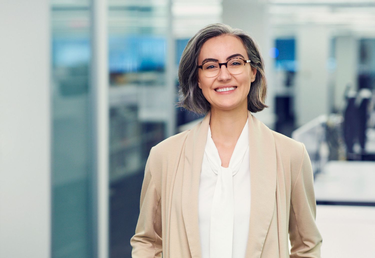 Smiling businesswoman with glasses and short gray hair wearing a beige blazer in a modern office.