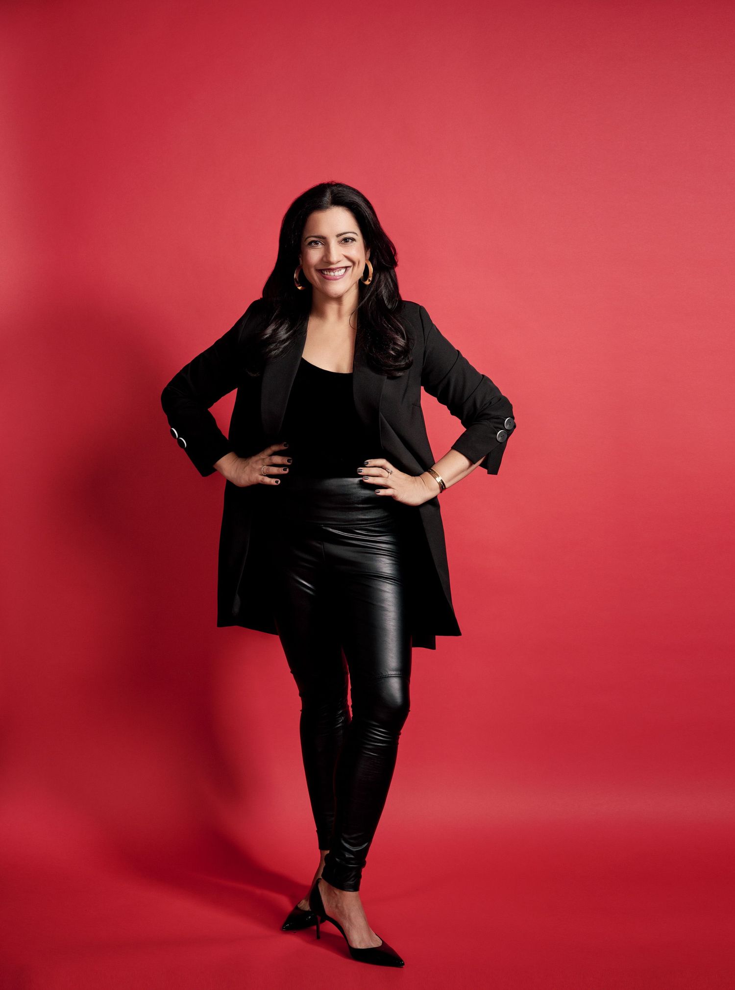 Woman in black blazer and leather pants posing confidently against a bold red background.