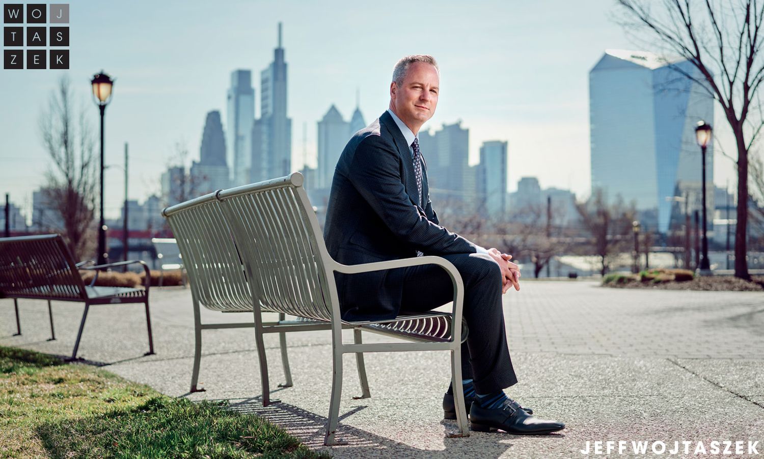 Man in suit sitting on bench with Philadelphia skyline in background on clear day.