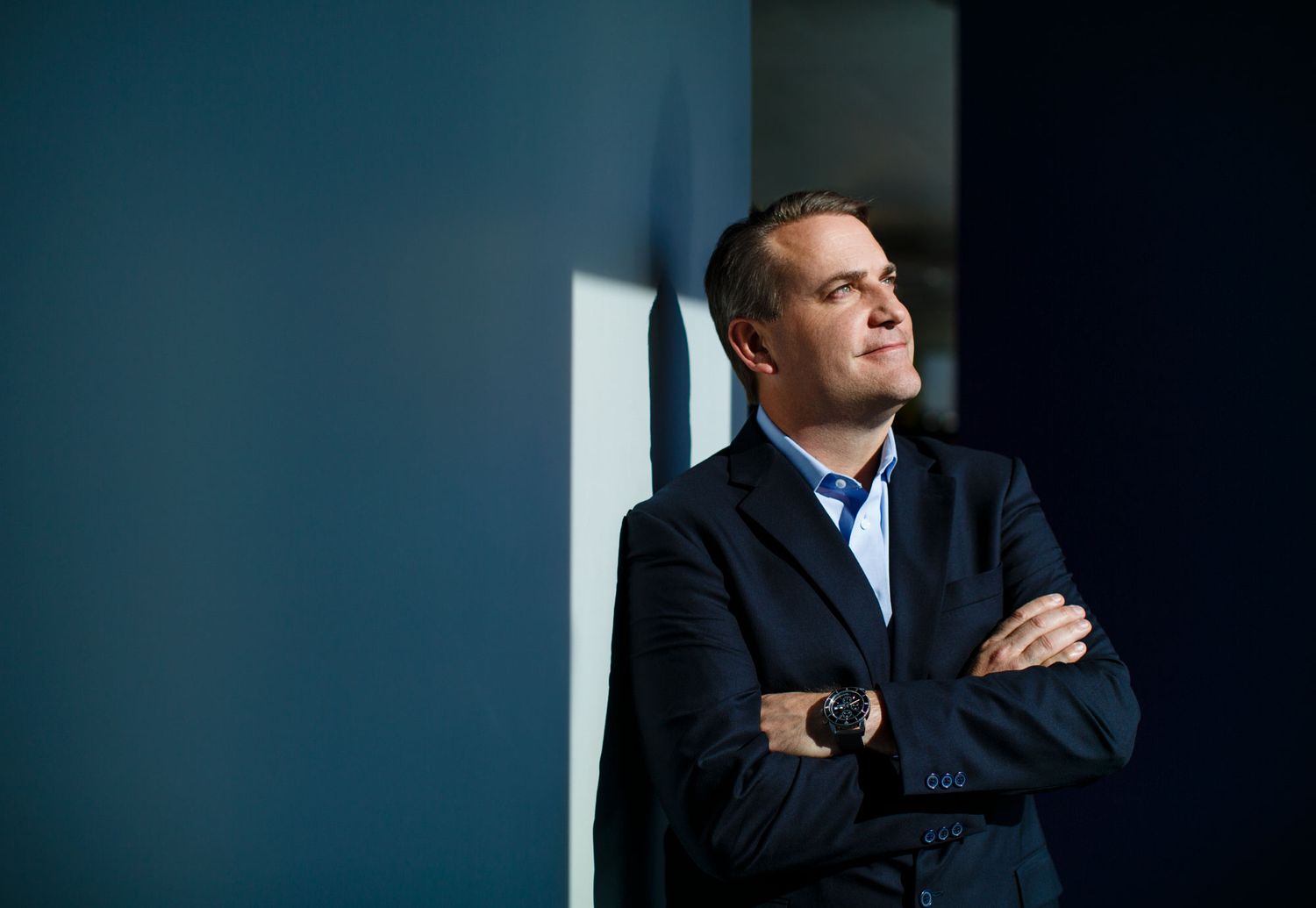 Confident businessman in a navy blazer leaning against a blue wall with arms crossed, looking up.