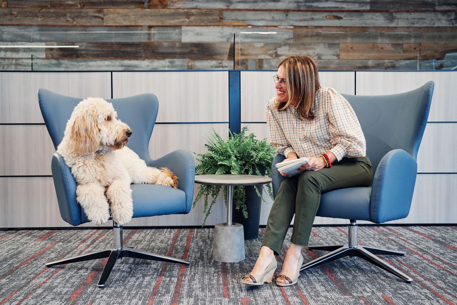Woman sitting in modern office lounge chair smiling at fluffy dog seated beside her.