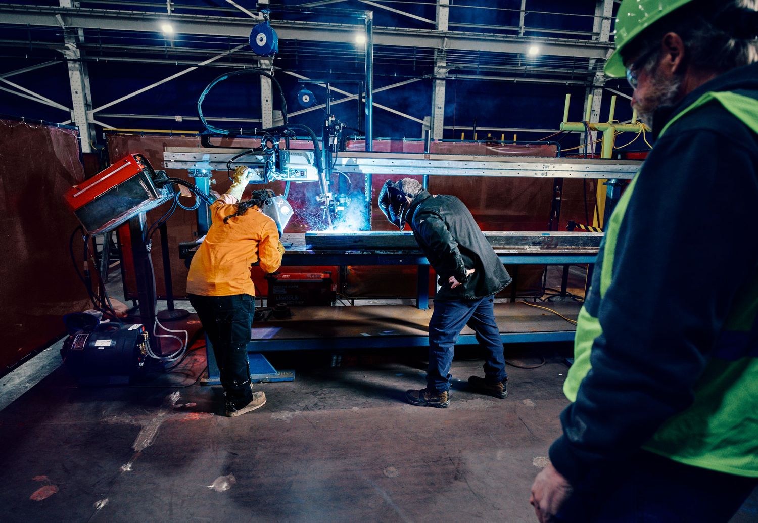 Workers in safety gear operating automated welding machine on large steel panel in warehouse.