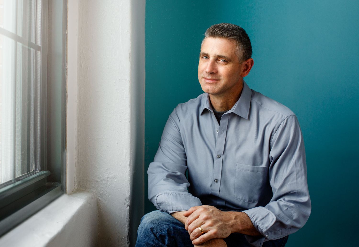 Thoughtful man in casual blue shirt sits by window in teal-walled room with natural light.