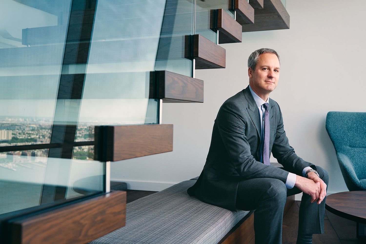Professional businessman in suit sitting on bench in modern office with glass staircase and city view.