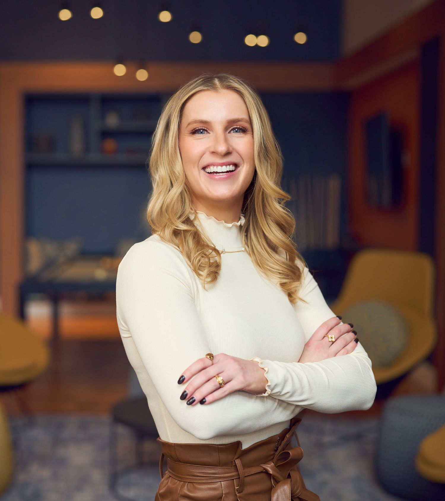 Smiling blonde woman in white turtleneck and brown pants poses with arms crossed indoors.