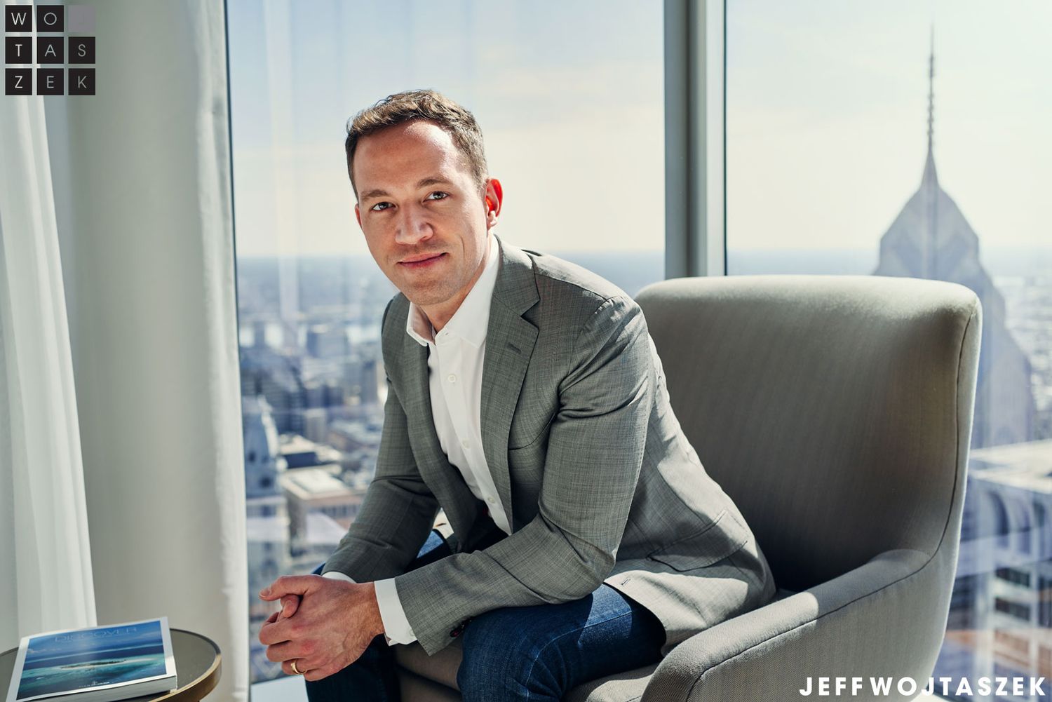 Professional man in gray blazer seated in modern chair with city skyline visible through large window behind him.