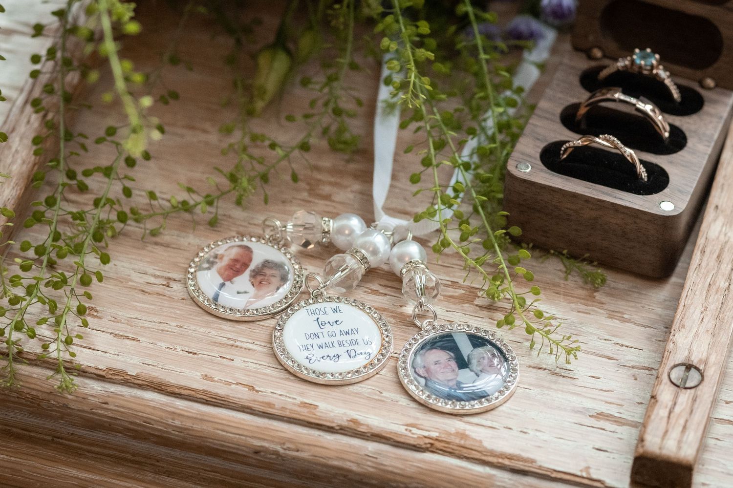 Wedding keepsake charms with photos displayed on a wooden tray beside rings and greenery.