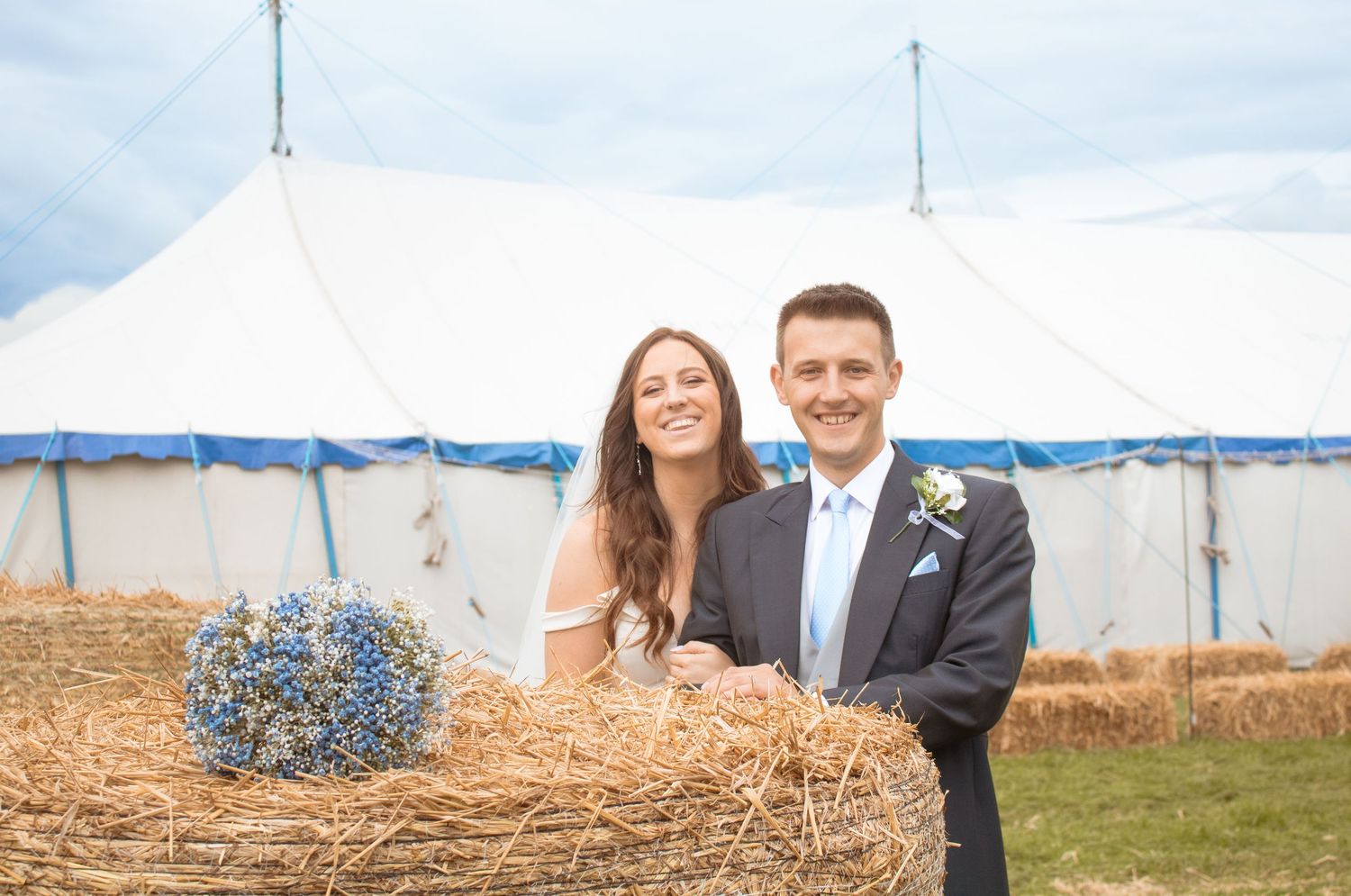 Happy bride and groom pose beside hay bales with blue bouquet in front of white marquee tent.
