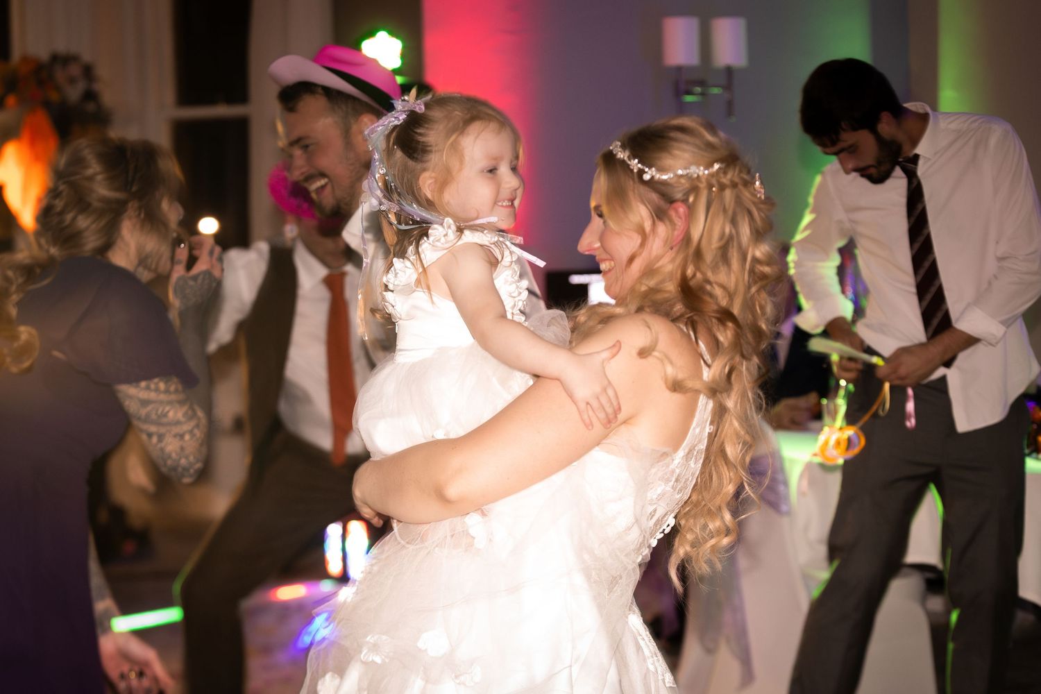 Bride in white gown dances with young flower girl at lively wedding reception with colorful lights and guests.