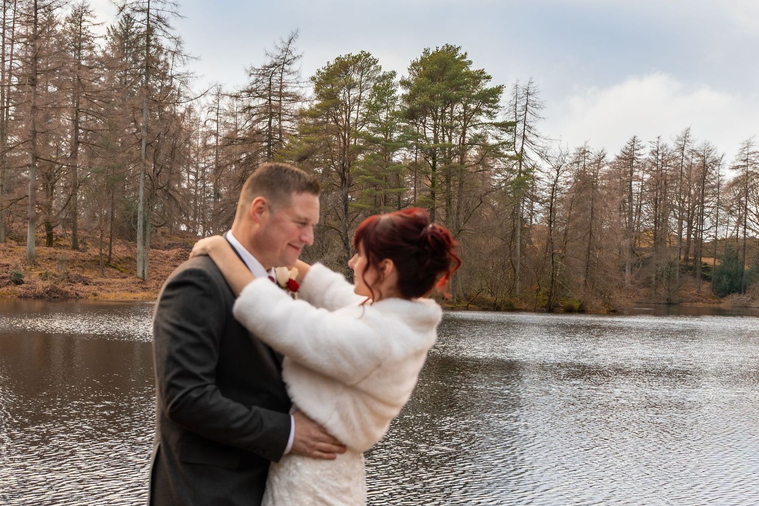 Bride and groom embrace romantically by a serene lake surrounded by autumn trees on their wedding day.