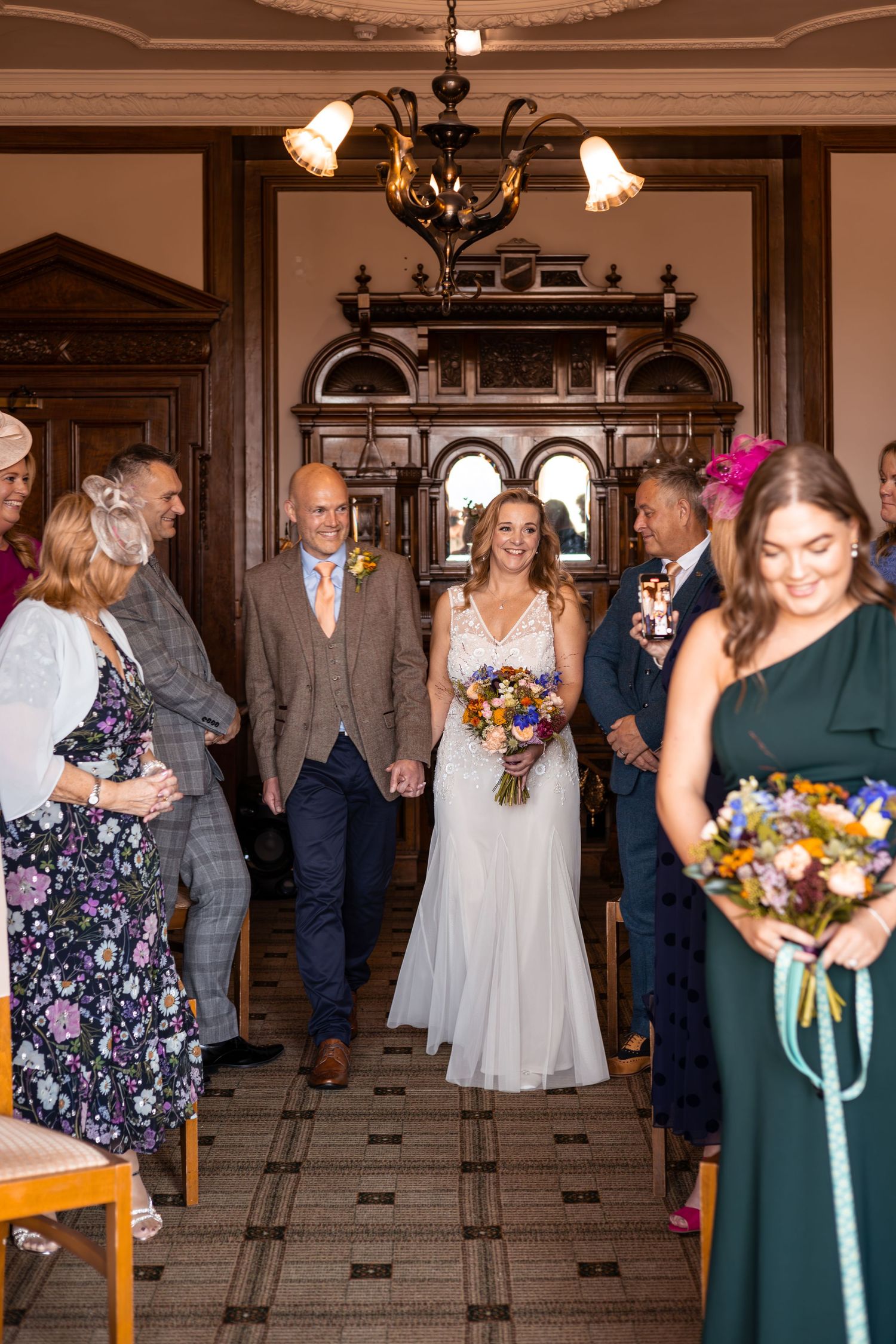 Bride and groom walk down the aisle hand in hand, surrounded by smiling guests in an elegant wood-paneled venue.