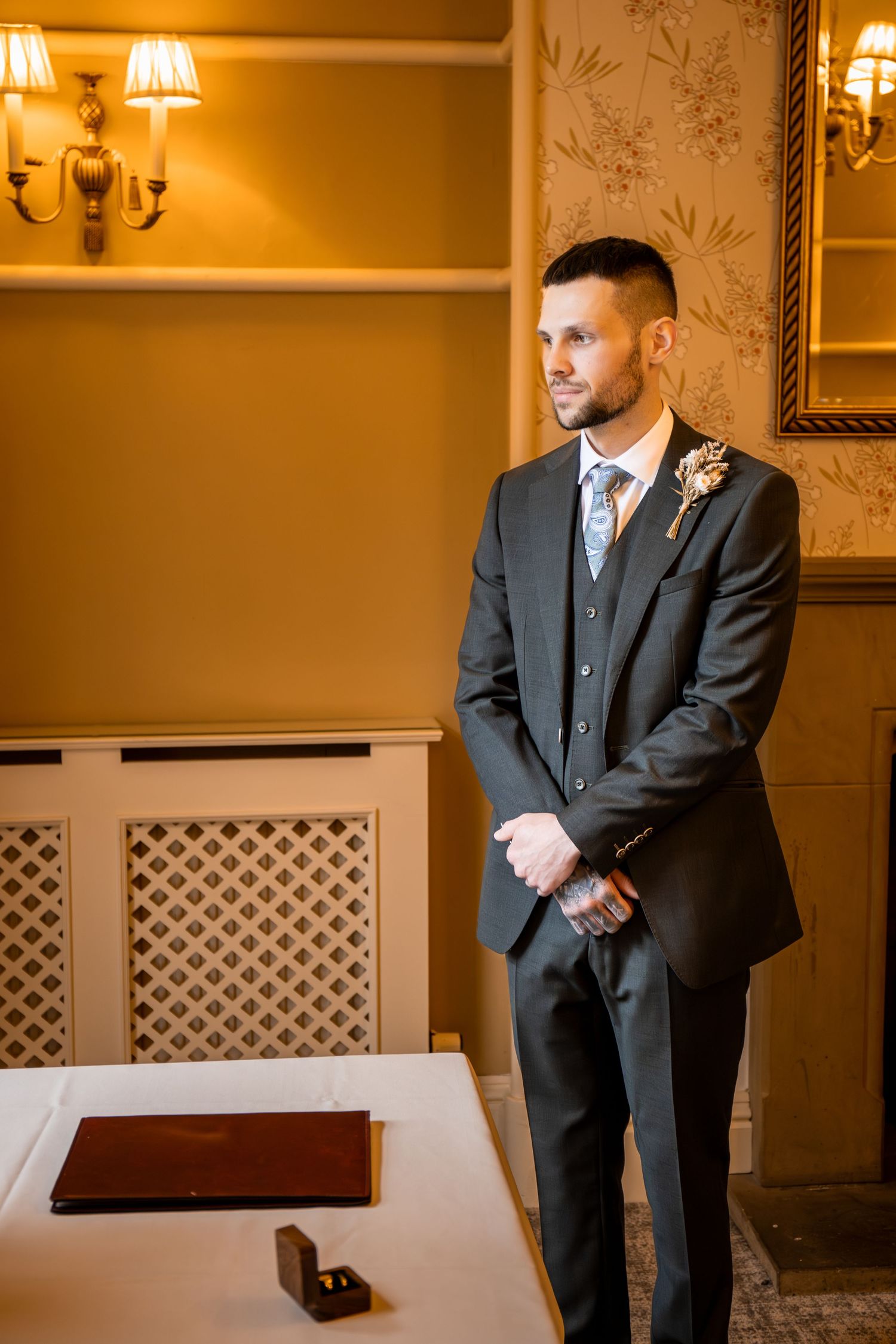 Groom in dark suit with boutonniere stands by table with wedding rings in elegant warmly lit room.