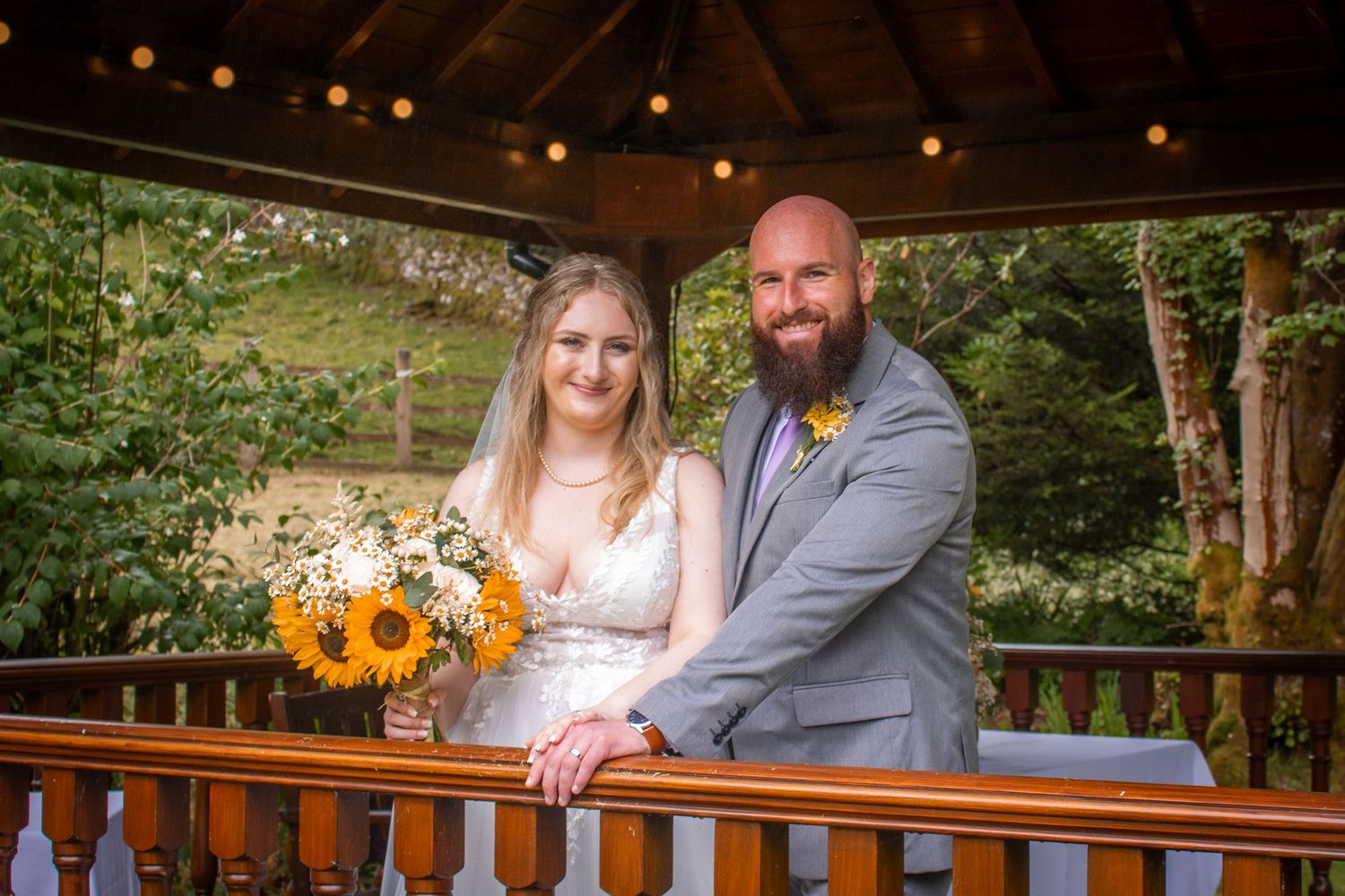 Bride and groom smiling on a wooden gazebo, bride holding sunflower bouquet, groom in gray suit, lush green backdrop.